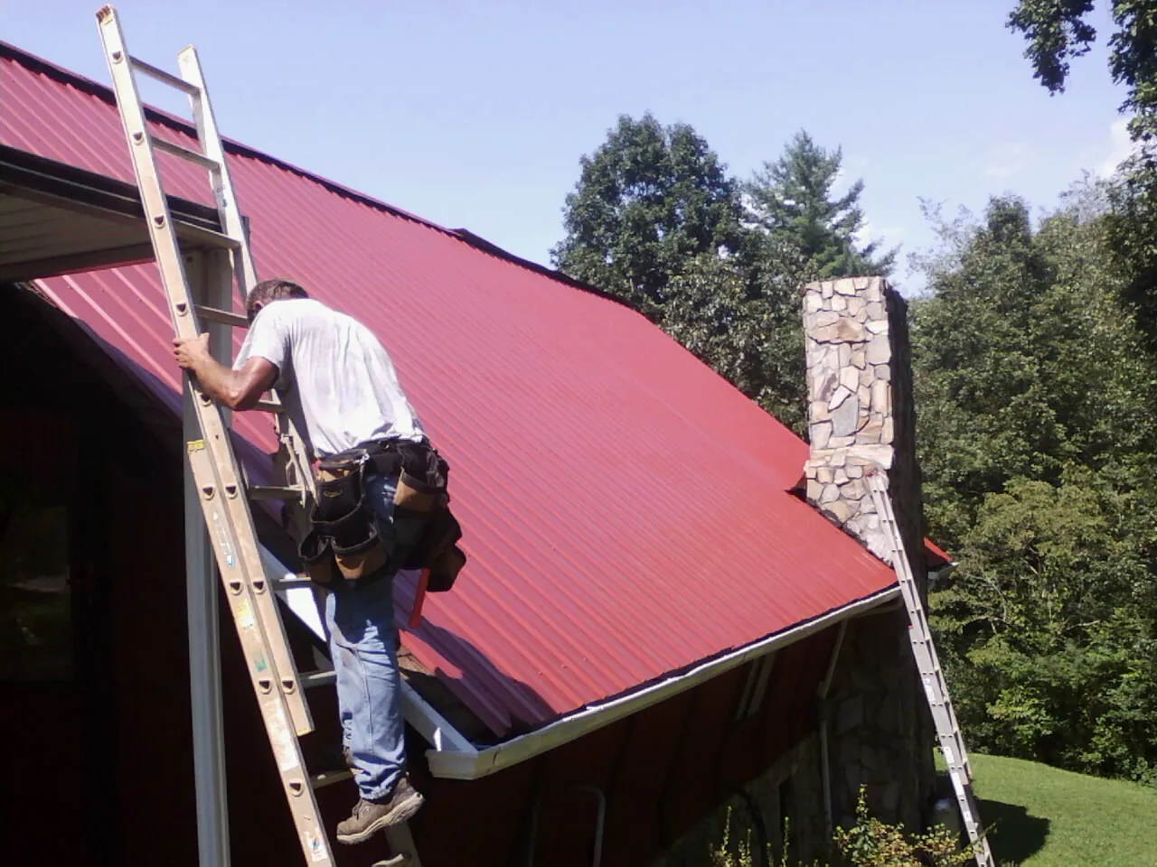 Roofer on a ladder beside a red metal roof and stone chimney — Soffit & Fascia Repair by Roofing Brothers in Northpoint